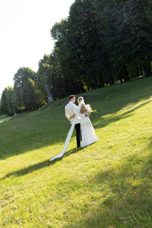 Das Brautpaar steht in der Morgensonne nebeneinander im Schlosspark. Der Anzug ist klassisch.