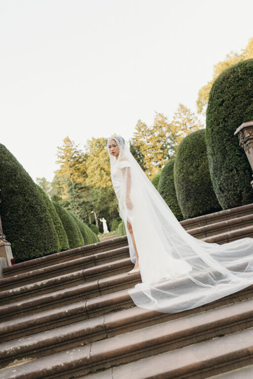 Die Braut trägt ein Brautkleid sowie einen langen Schleier, sie steht auf der Treppe und scheint in die Kamera der Hochzeitsfotografin aus Hamburg Laura Kaiser.