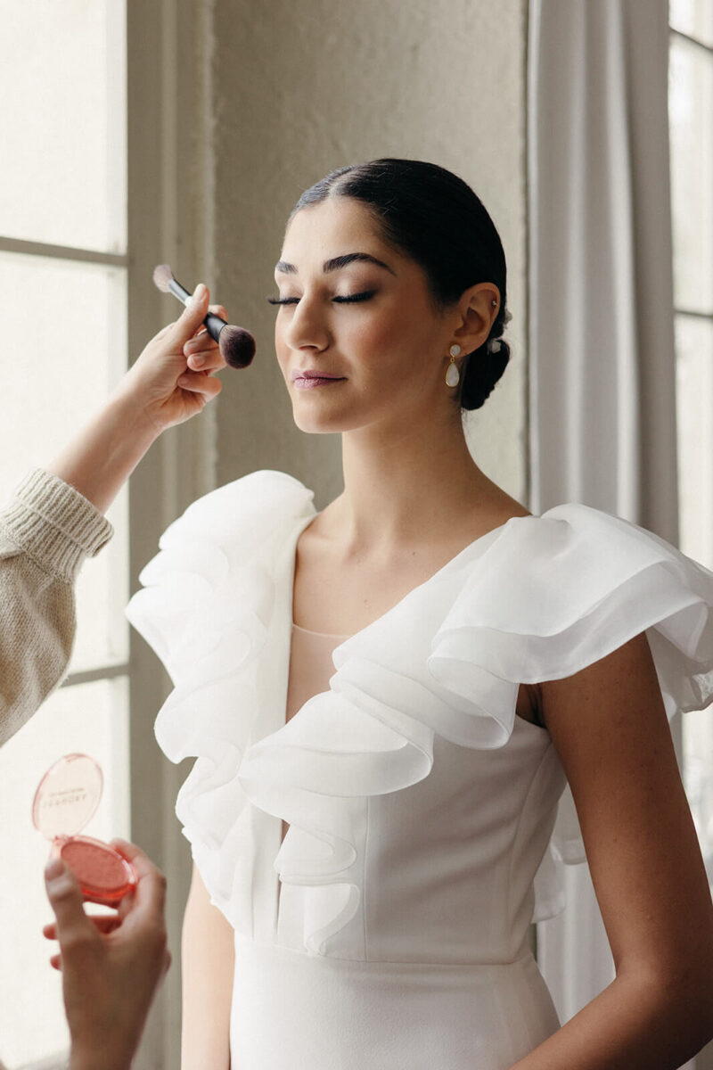 Die Braut wird vor dem Fenster stehend geschminkt.